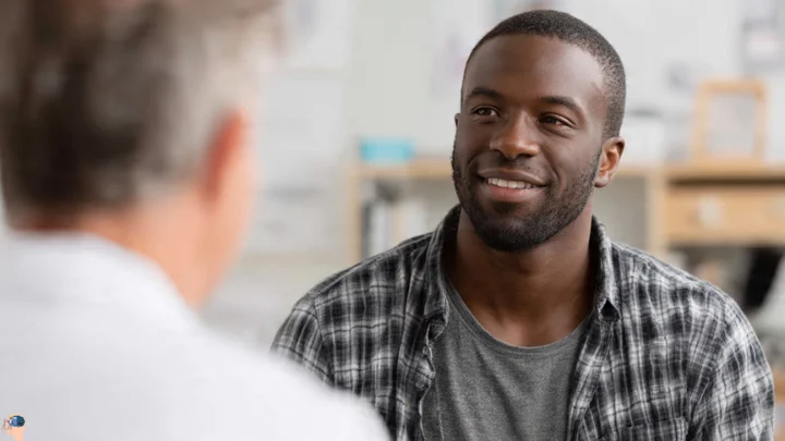 A smiling Black man with a beard looking at a dermatologist, discussing skin care for black men in a clinic or office setting.