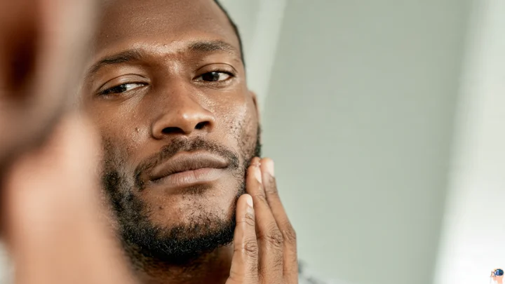 A close-up image of a Black man examining the texture of his face and short beard in a mirror, focusing on his daily black men skin care needs.